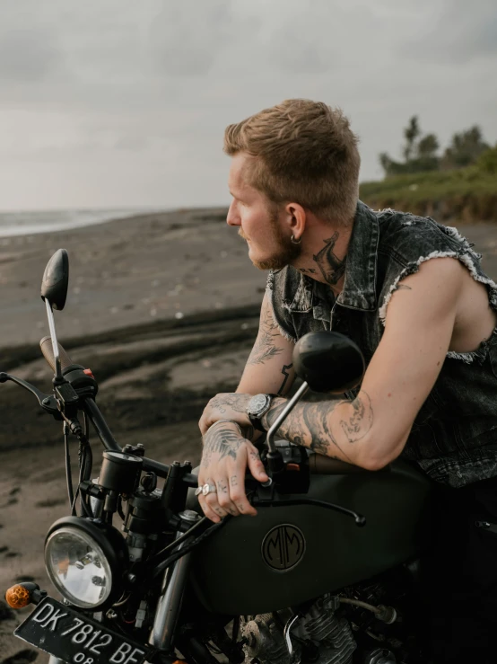 a man sitting on a motorcycle on a beach, a tattoo, trending on pexels, wearing a vest, non binary model, black sand, profile image