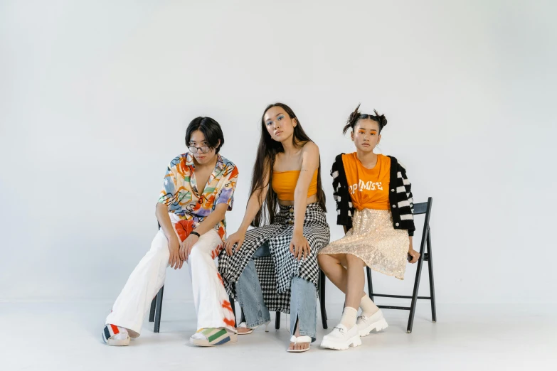 three women sitting next to each other on a chair, an album cover, trending on pexels, asian female, colourful clothes, tilt and orange, in a white room