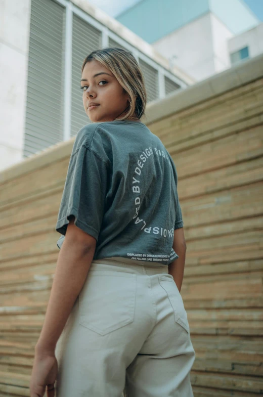 a woman standing in front of a wooden wall, an album cover, by Christen Dalsgaard, trending on unsplash, aestheticism, wearing pants and a t-shirt, in white lettering, dressed in a gray, back view »