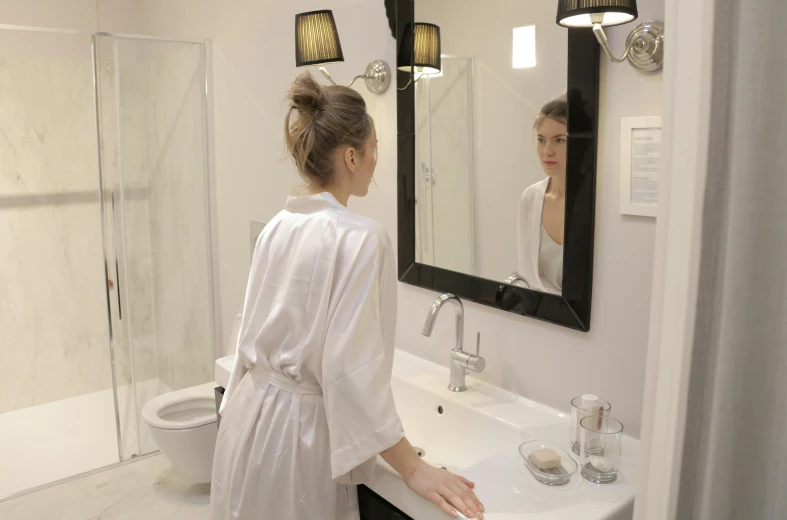 a woman in a bathrobe stands in a white bathroom looking at herself in the mirror