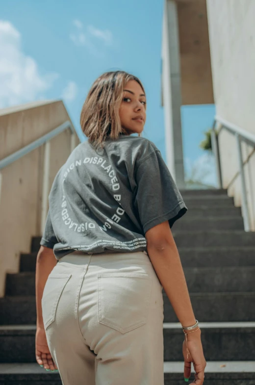 a woman walking up a flight of stairs, an album cover, trending on unsplash, wearing pants and a t-shirt, curvy accentuated booty, dressed in a gray, in white lettering