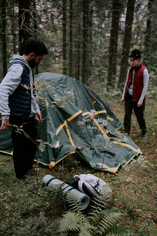 a man standing next to a tent in the woods, an album cover, by Attila Meszlenyi, unsplash, two male, behind the scenes photo, injured, ( ( theatrical ) )