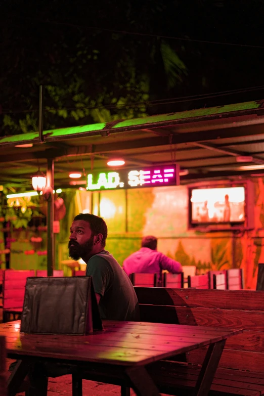 a man sitting at a table outside of a restaurant, inspired by Nan Goldin, unsplash, jamaican colors, laos, neon bar lights, sittin