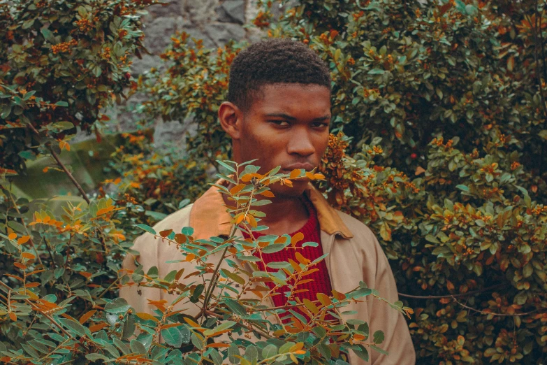 a man standing in front of a bush with a flower in his mouth, by Maxwell Bates, pexels contest winner, brown skinned, green and brown clothes, male teenager, looking serious