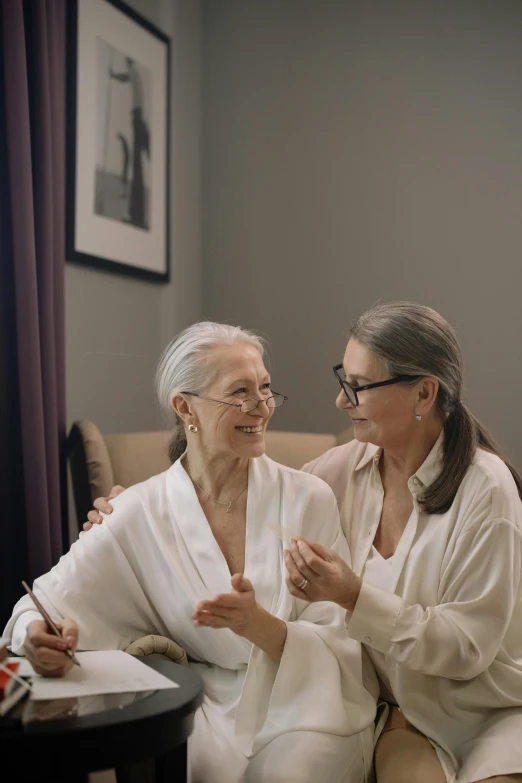 a couple of women sitting on top of a couch, wearing a grey robe, elderly, wearing white suit and glasses, sitting in bedroom