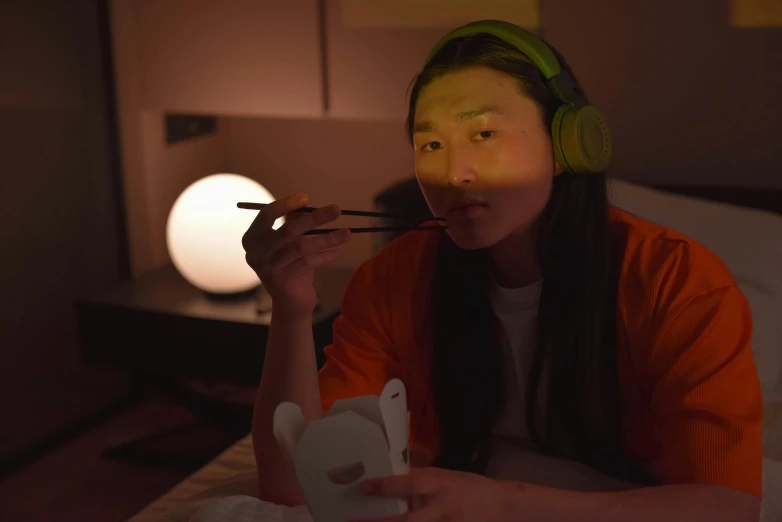 a woman sitting on a bed with a pair of chopsticks in her mouth, inspired by Liam Wong, glowing oled visor, wearing cat ear headphones, ikea, tea drinking and paper lanterns