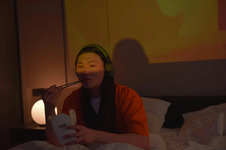 a woman sitting on top of a bed next to a lamp, inspired by Zhang Xiaogang, video art, headphones on head, eating, orange lighting, asian face