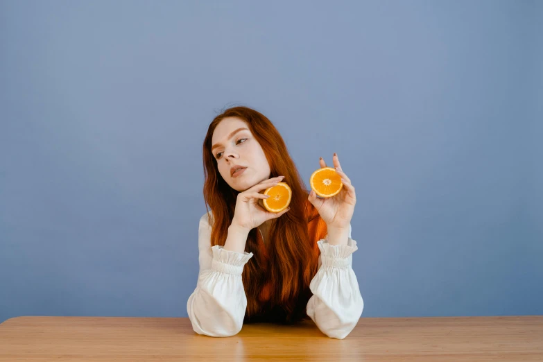 a woman sitting at a table with an orange slice in front of her face, an album cover, by Gavin Hamilton, pexels contest winner, she has long redorange hair, posing with crossed arms, sentient fruit, amber and blue color scheme