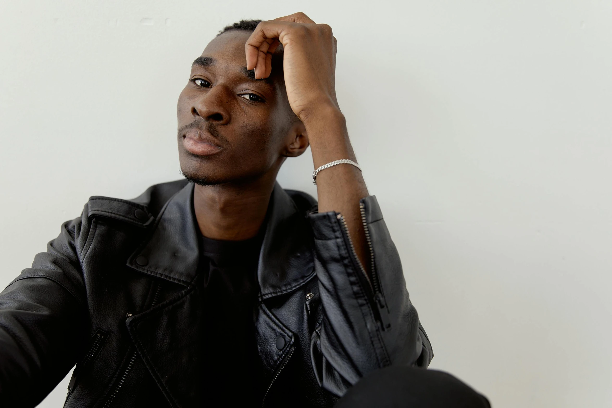 a man sitting in front of a white wall, an album cover, inspired by Theo Constanté, trending on pexels, wearing a black leather jacket, adut akech, thoughtful pose, portrait featured on unsplash