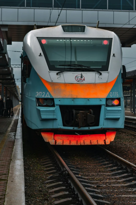 a blue and orange train pulling into a train station, by Attila Meszlenyi, front facing the camera, square, high quality photo, thumbnail
