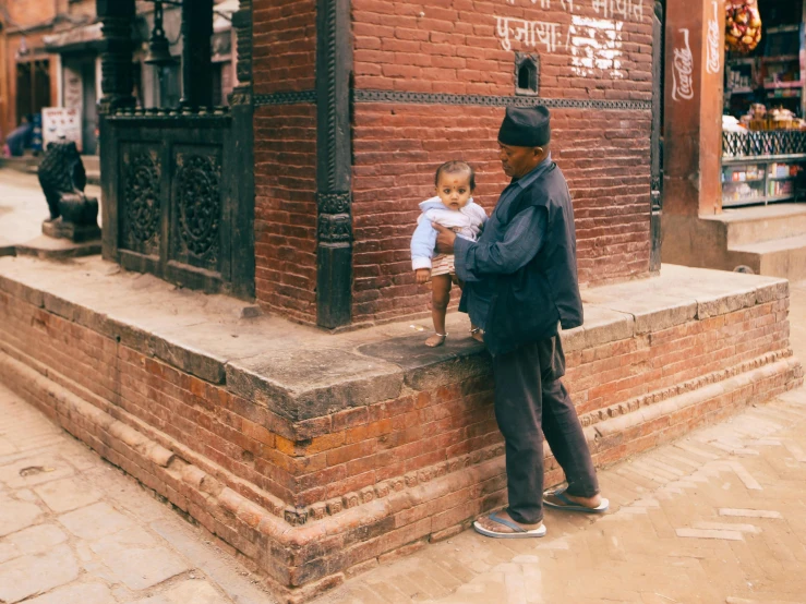 a man standing next to a brick building holding a baby, by Matija Jama, pexels contest winner, nepali architecture buildings, standing over a tomb stone, old cmputers on the sidewalk, hugging his knees