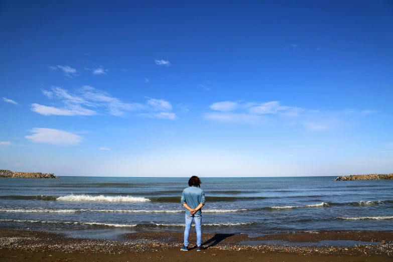 a woman standing on top of a beach next to the ocean, a picture, inspired by Storm Thorgerson, unsplash, renaissance, cloudless sky, handsome man, facing away from the camera, sparsely populated