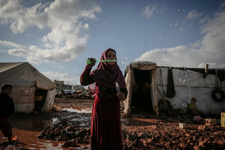 a woman standing in front of a group of tents, by Ibrahim Kodra, pexels contest winner, hurufiyya, bubbles in the air, damaged structures, holding flask in hand, refugees