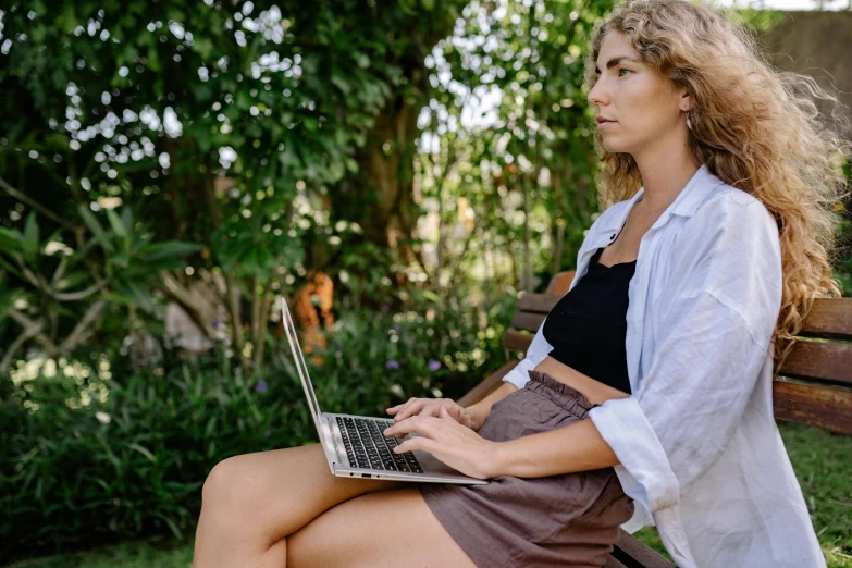 a woman sitting on a bench using a laptop, a portrait, by Alice Mason, unsplash, happening, in a jungle, slightly tanned, thigh focus, looking across the shoulder