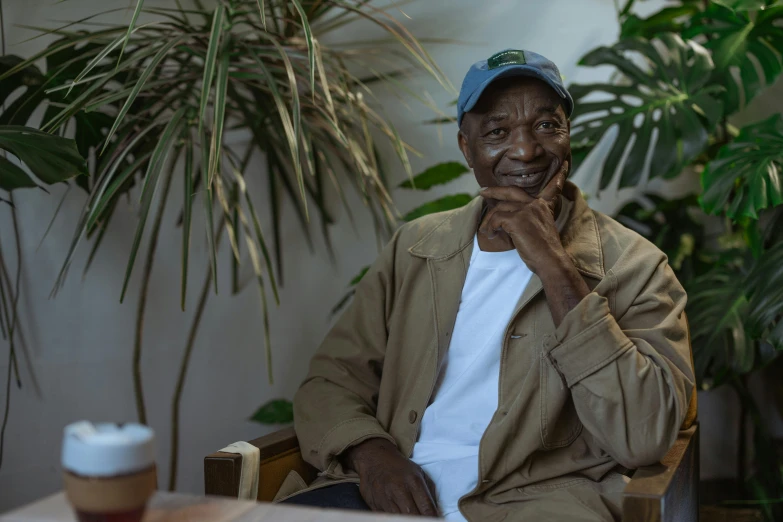 an older man with wrinkles sits in front of potted palm trees