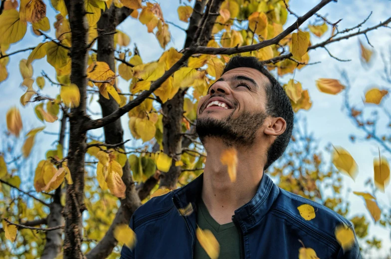 a man standing in front of a tree with yellow leaves, pexels contest winner, mutahar laughing, young greek man, avatar image, low - angle shot