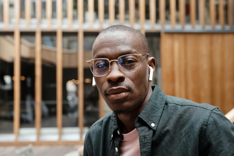 a man wearing glasses standing in front of a building, inspired by Barthélemy Menn, pexels contest winner, maria borges, shaven, ignant, looking towards camera