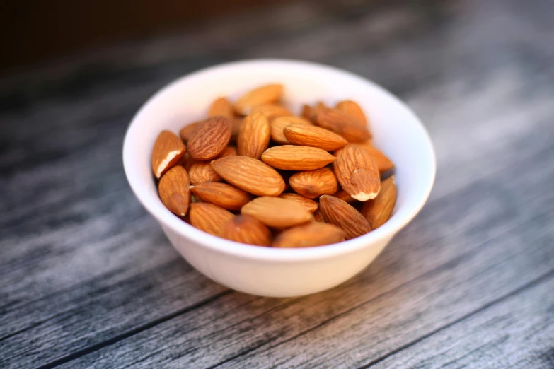 a white bowl filled with almonds on top of a wooden table, by Carey Morris, pexels, 🦩🪐🐞👩🏻🦳, snacks, close-up photo, bowl filled with food