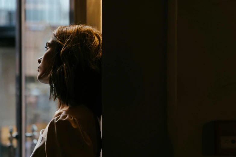 a woman standing in front of a window looking out, a picture, inspired by Elsa Bleda, trending on pexels, emma watson with anxious, woman very tired, in the evening, side profile shot