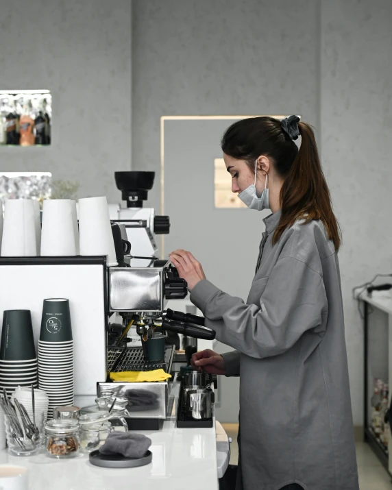 a woman wearing a face mask working in a coffee shop, by Emma Andijewska, pexels contest winner, process art, coffee machine, wearing a grey robe, wearing rr diner uniform, detailed information