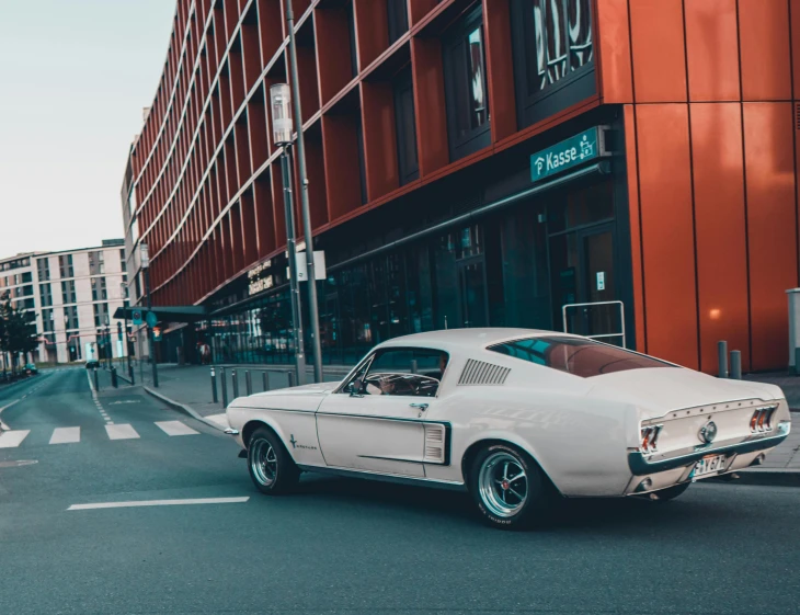 a white car driving down a street next to tall buildings, by Adam Marczyński, pexels contest winner, modernism, green 1967 ford mustang gt, restomod, norway, 🚿🗝📝