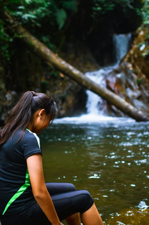 a woman sitting on a rock in front of a waterfall, sumatraism, looking sad, doing a prayer, next to a small river