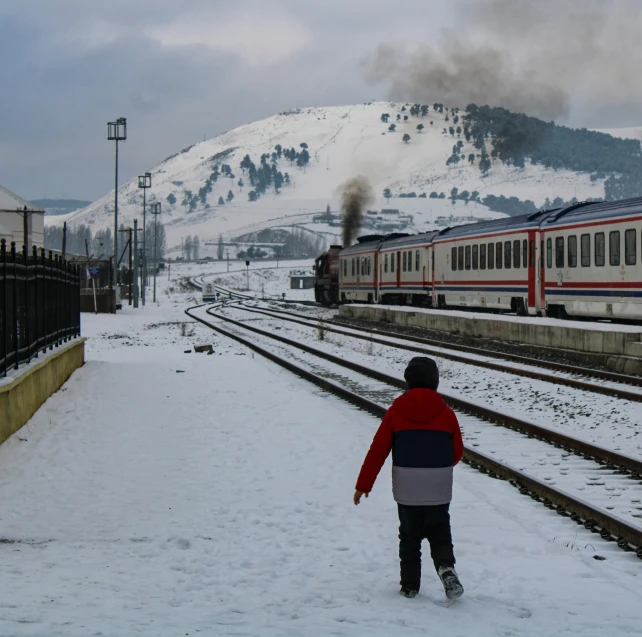 a person walking in the snow next to a train, by Lucia Peka, pexels contest winner, little boy, hills in the background, square, platform