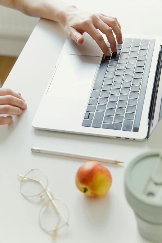 a close up of a person typing on a laptop, having a snack, holding an apple, white table, multiple stories