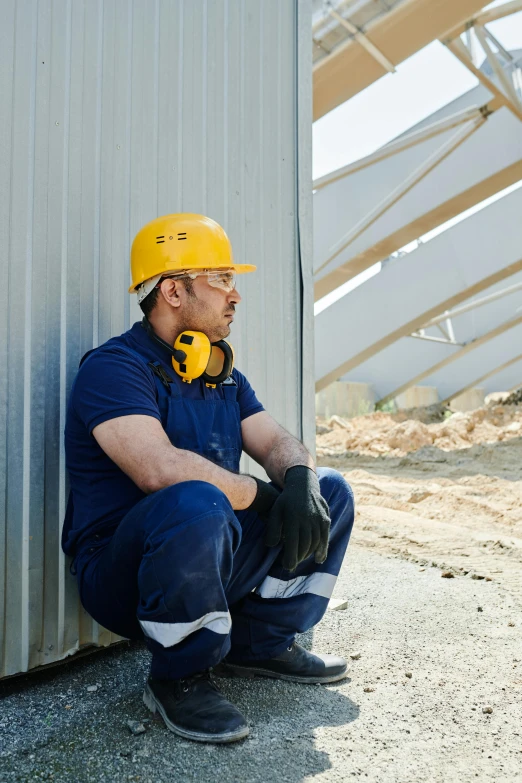 a man sitting on the ground in front of a building, yellow helmet, profile image, calmly conversing 8k, sleek metal ears