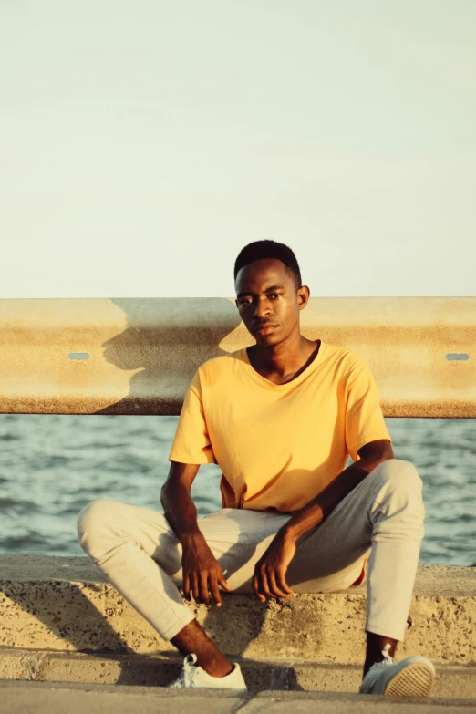 a man sitting on a ledge next to a body of water, wearing yellow croptop, dark complexion, androgynous person, oceanside