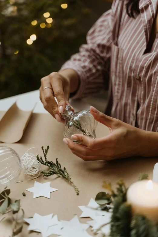 a woman sitting at a table in front of a christmas tree, crafting, glass bulbs, sleek hands, botanical