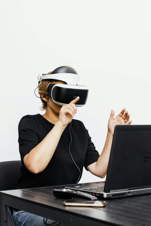 a woman sitting at a table in front of a laptop wearing a virtual reality headset, inspired by Li Di, slightly minimal, press shot, eka's portal, emotion