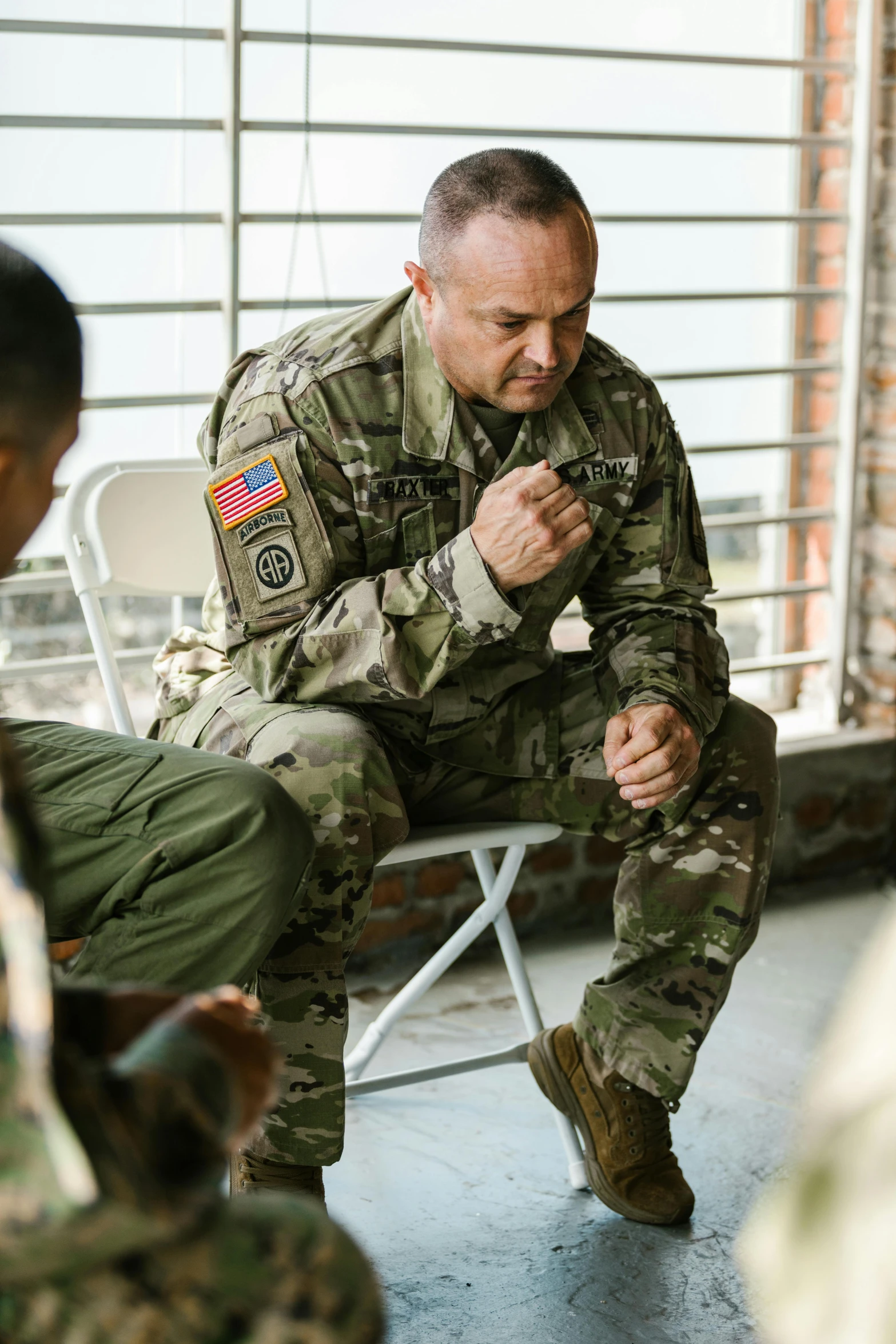 a soldier sitting on a bench looking at his cell phone, sitting with wrists together, pray, a man, sitting across the room