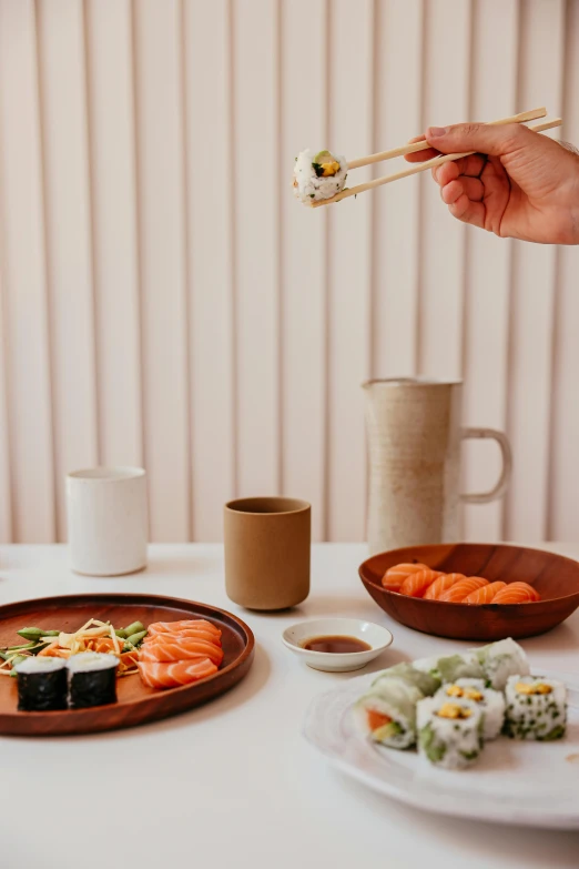 a person holding chopsticks over a plate of sushi, inspired by Tōshi Yoshida, wood cups, curated collections, on kitchen table, minimalistic design