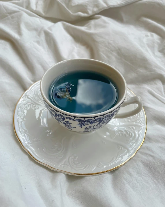 a cup of tea sitting on top of a saucer, laying on a bed, with blue skin, ☁🌪🌙👩🏾, blue and green water