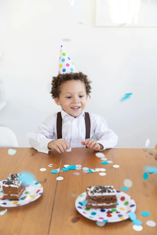 a little boy that is sitting at a table, confetti, white with chocolate brown spots, wearing a party hat, curated collections