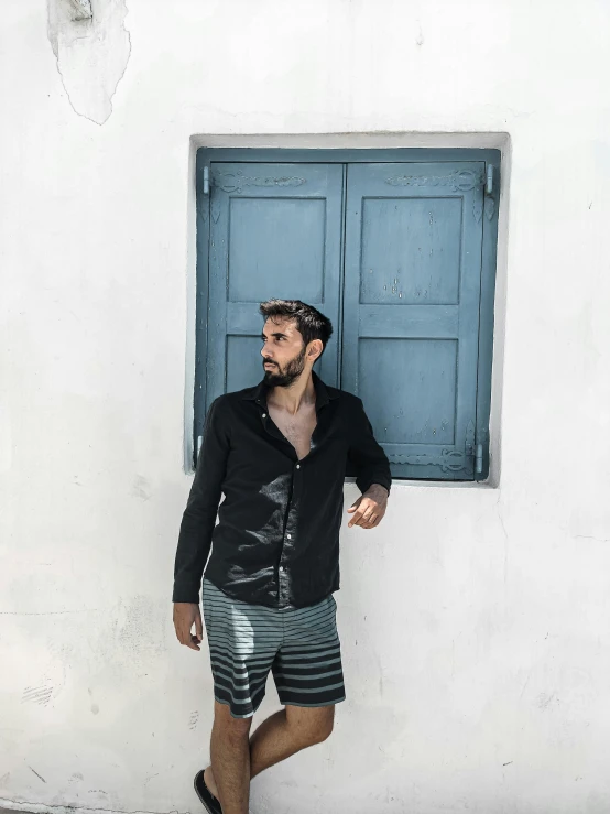 a man standing in front of a blue window, a black and white photo, inspired by Constantine Andreou, wearing black shorts, wearing stripe shirt, in the design of eliot kohek, instagram photo shoot