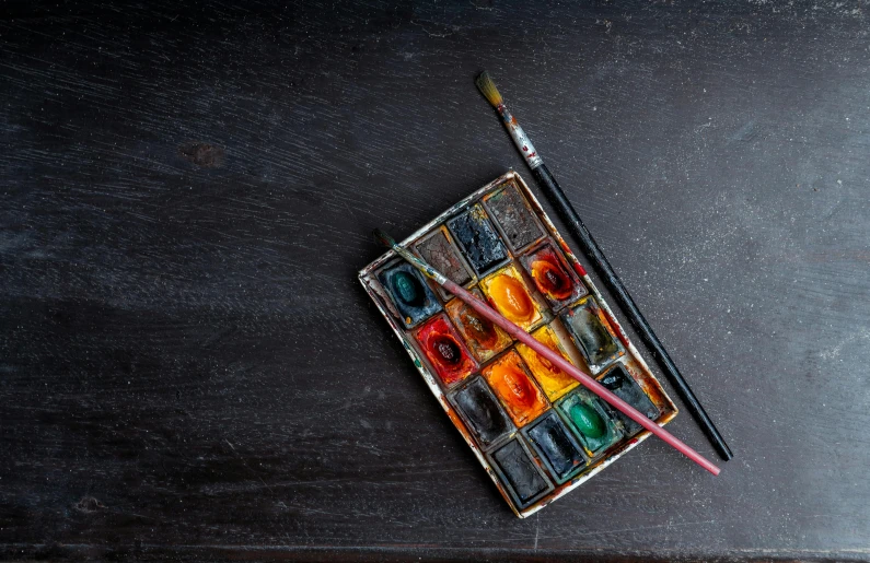 a box of watercolors sitting on top of a wooden table, inspired by artist, pexels contest winner, black studio background color, paintbrush, rectangle, grungy; colorful