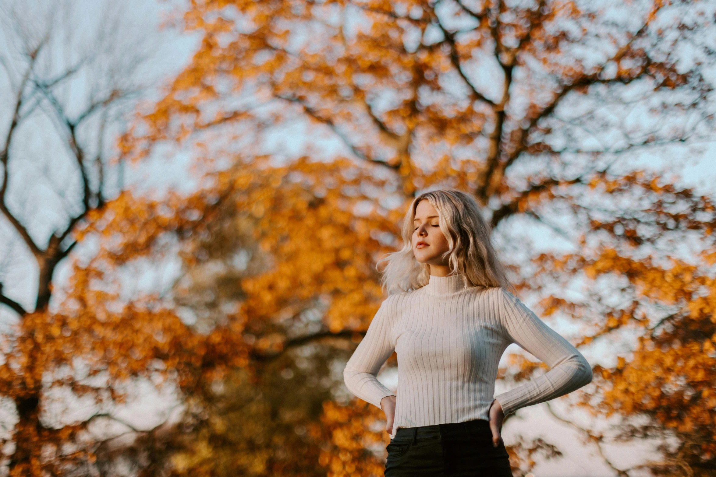 a woman standing in front of a tree with her hands on her hips, by Emma Andijewska, pexels contest winner, in white turtleneck shirt, autumn light, erin moriarty, white and orange