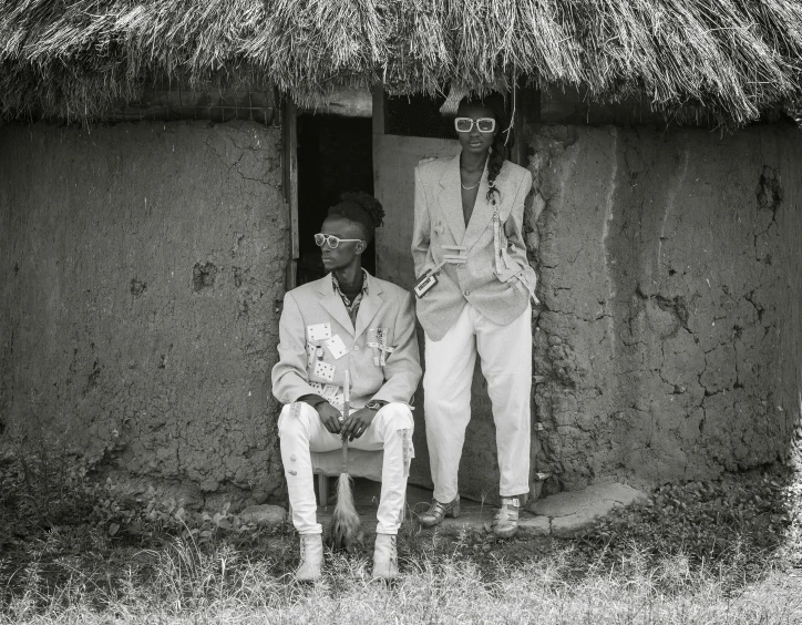 a couple of men sitting in front of a hut, a black and white photo, by Maurycy Gottlieb, pexels contest winner, afrofuturism, wearing white suit and glasses, portrait of tall, unmistakably kenyan, young thug