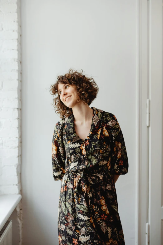 a woman standing in front of a window, an album cover, by Nina Hamnett, dark short curly hair smiling, dressed in a flower dress, jovana rikalo, full frame image