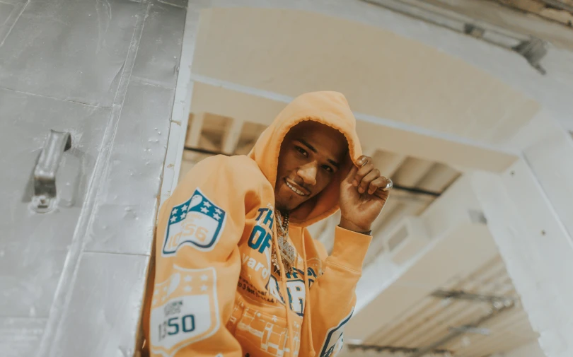 a man in a yellow hoodie leaning against a wall, rapper jewelry, orange and white, mecca, 2 k aesthetic