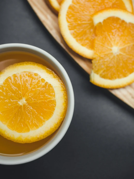 a bowl filled with orange slices next to a cutting board, trending on unsplash, is ((drinking a cup of tea)), bottom body close up, thumbnail, on a gray background