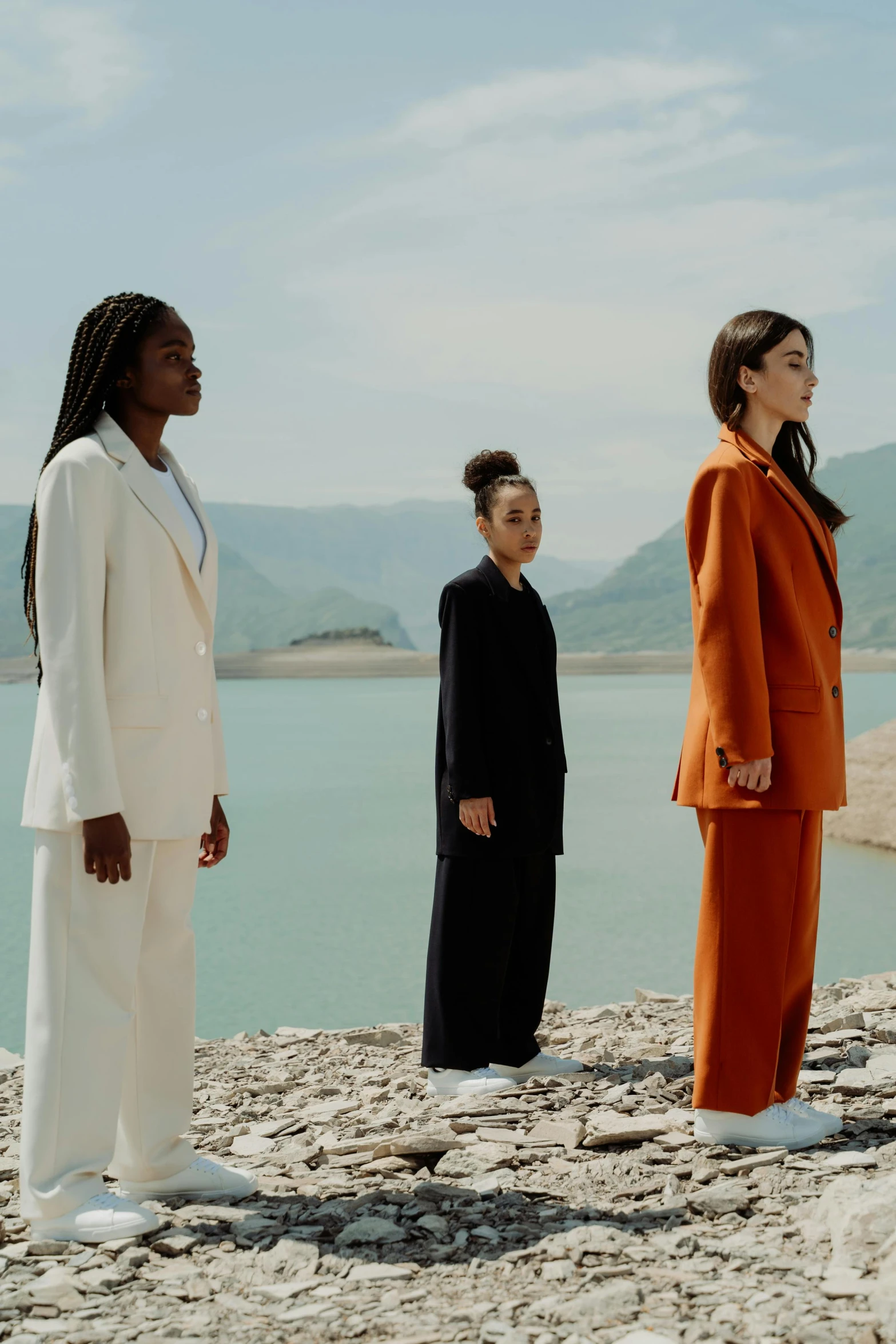 a group of people standing in front of a body of water, an album cover, inspired by Vanessa Beecroft, pexels contest winner, modernism, three piece suit, standing in front of a mountain, 3 young and beautiful women, diverse colors