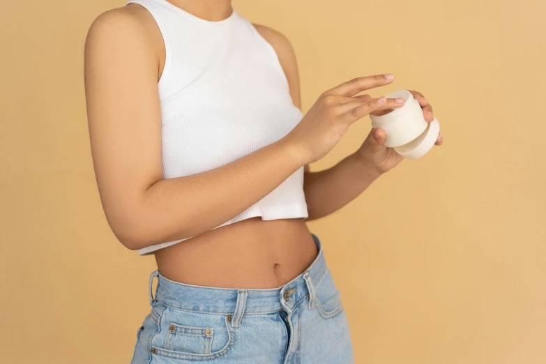 a woman in a white tank top holding a cup, trending on pexels, belly button showing, skincare, detailed product image, bandage on arms