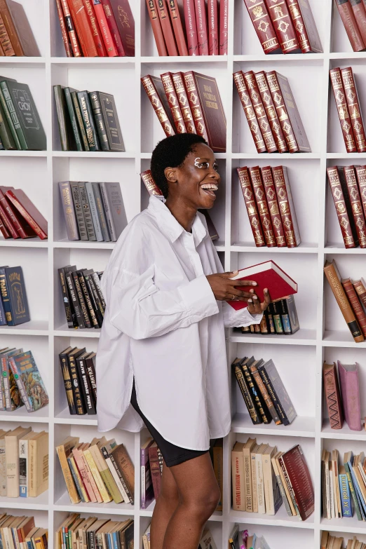 a woman standing in front of a bookshelf holding a book, happening, wearing a white button up shirt, dark-skinned, having a great time, grand library