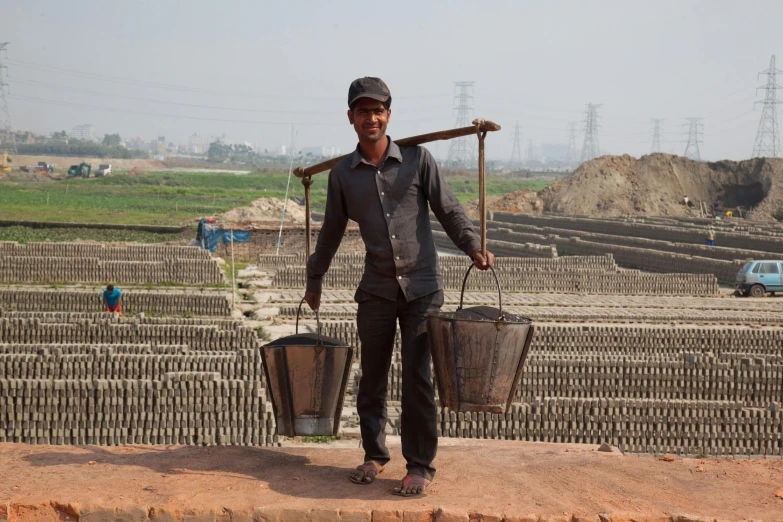 a man standing on top of a brick wall holding two buckets, by Jan Tengnagel, temporary art, bangladesh, iron smelting pits, 2 0 2 2 photo, small manufacture