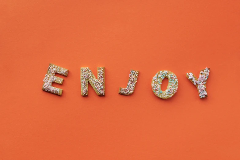 the word enjoy spelled in glitter on an orange background, by Emma Andijewska, trending on unsplash, letterism, cookies, covered in sprinkles, joyous wide memorable, eating cakes