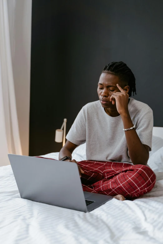 a man sitting on a bed using a laptop, trending on pexels, disappointed, afro tech, non-binary, wearing a baggy pajamas