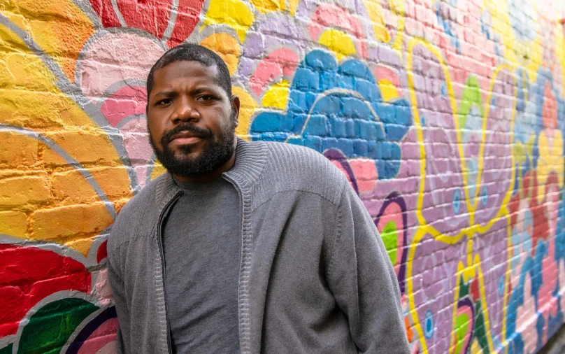 a man in a gray jacket leans against a brightly painted wall